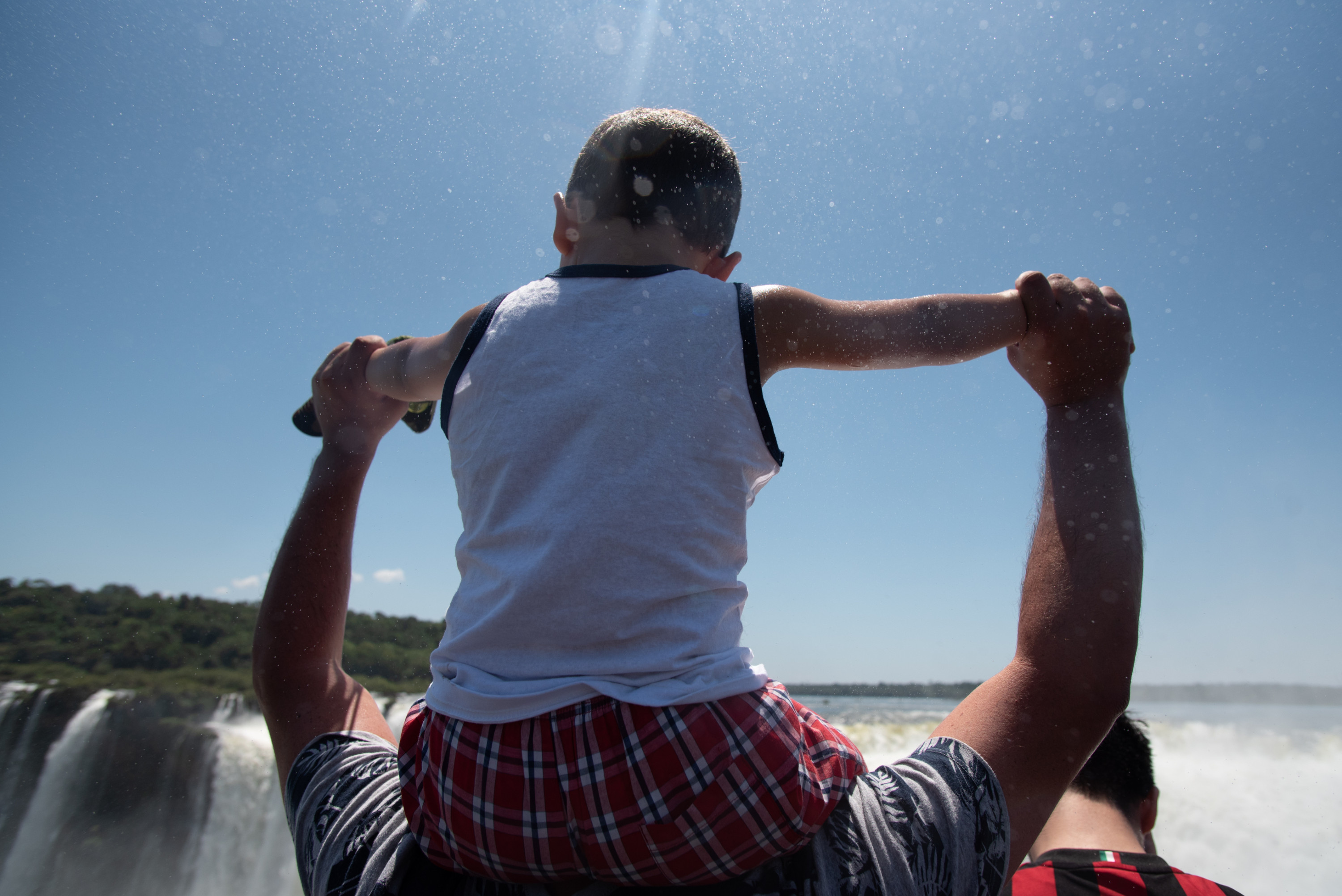 A small child sits on a person’s shoulders with its arms spread wide in the mist coming off of the waterfalls at the Devil’s Throat in Iguazú National Park.