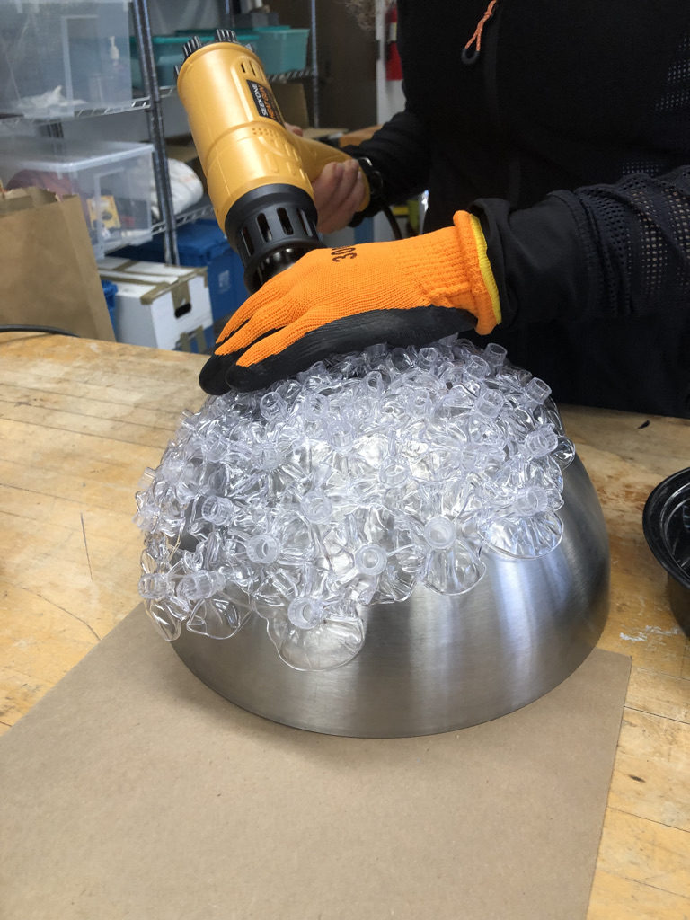 Ledón drills something into a large metal half-sphere that is affixed with ribbons of clear plastic. 