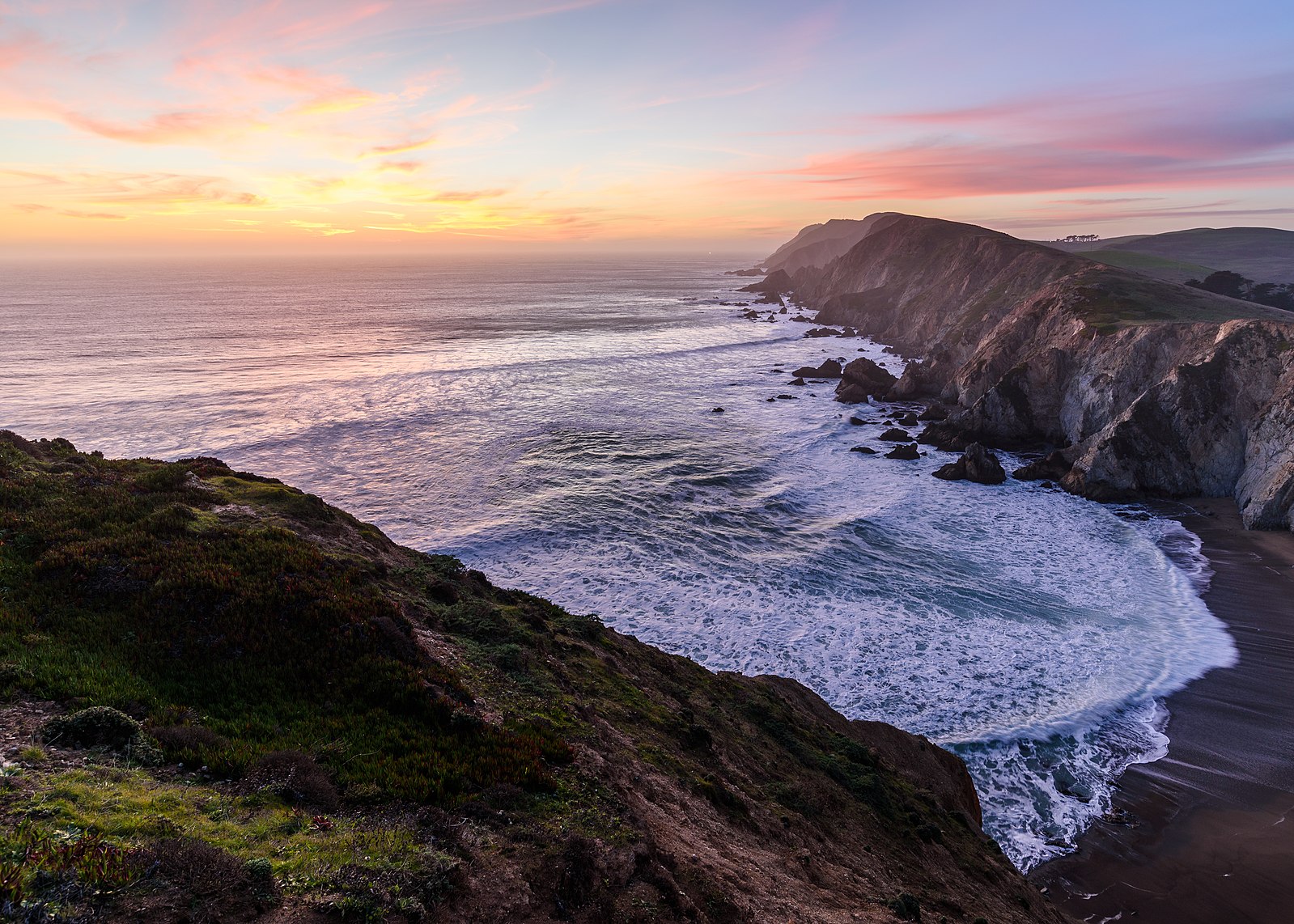 A small inlet at the Point Reyes National Seashore. Frothing ocean water pours into a bay area with rugged terrain.