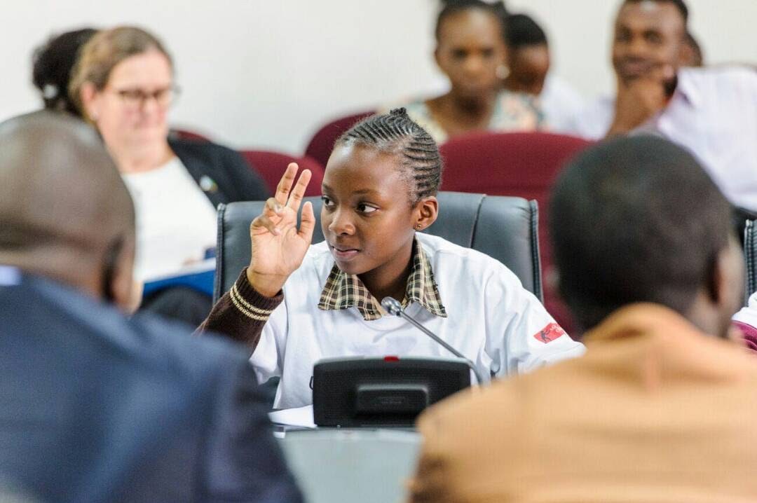 Mwangala sits at a conference table with several adults. She raises her hand in preparation to speak.