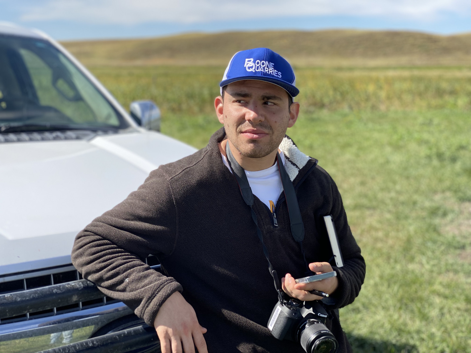 Adam leans against the hood of a truck and looks off into the distance with his DSLR camera hanging around his neck.