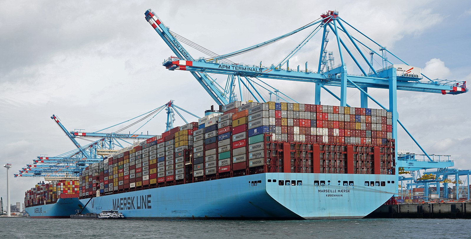 A large blue shipping vessel with many containers stacked on its deck rests at a dock with cranes towering above it.