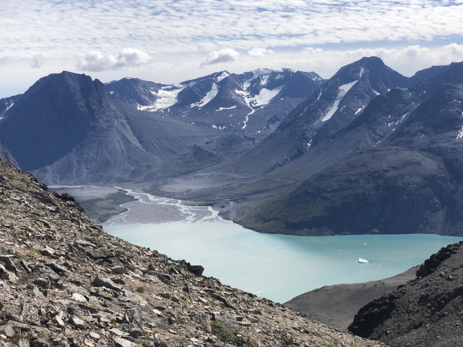 A mountainous landscape is in the distance, towering over a barren valley with a river running through it that opens up into a bright blue lake.