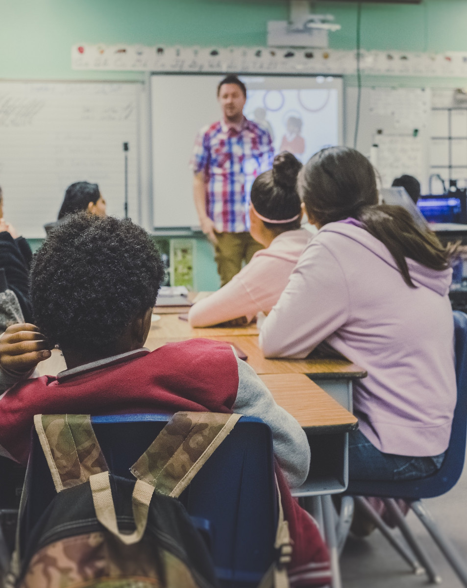 A group of teenagers sit at a table in a school classroom, looking towards an instructor standing at the white board.