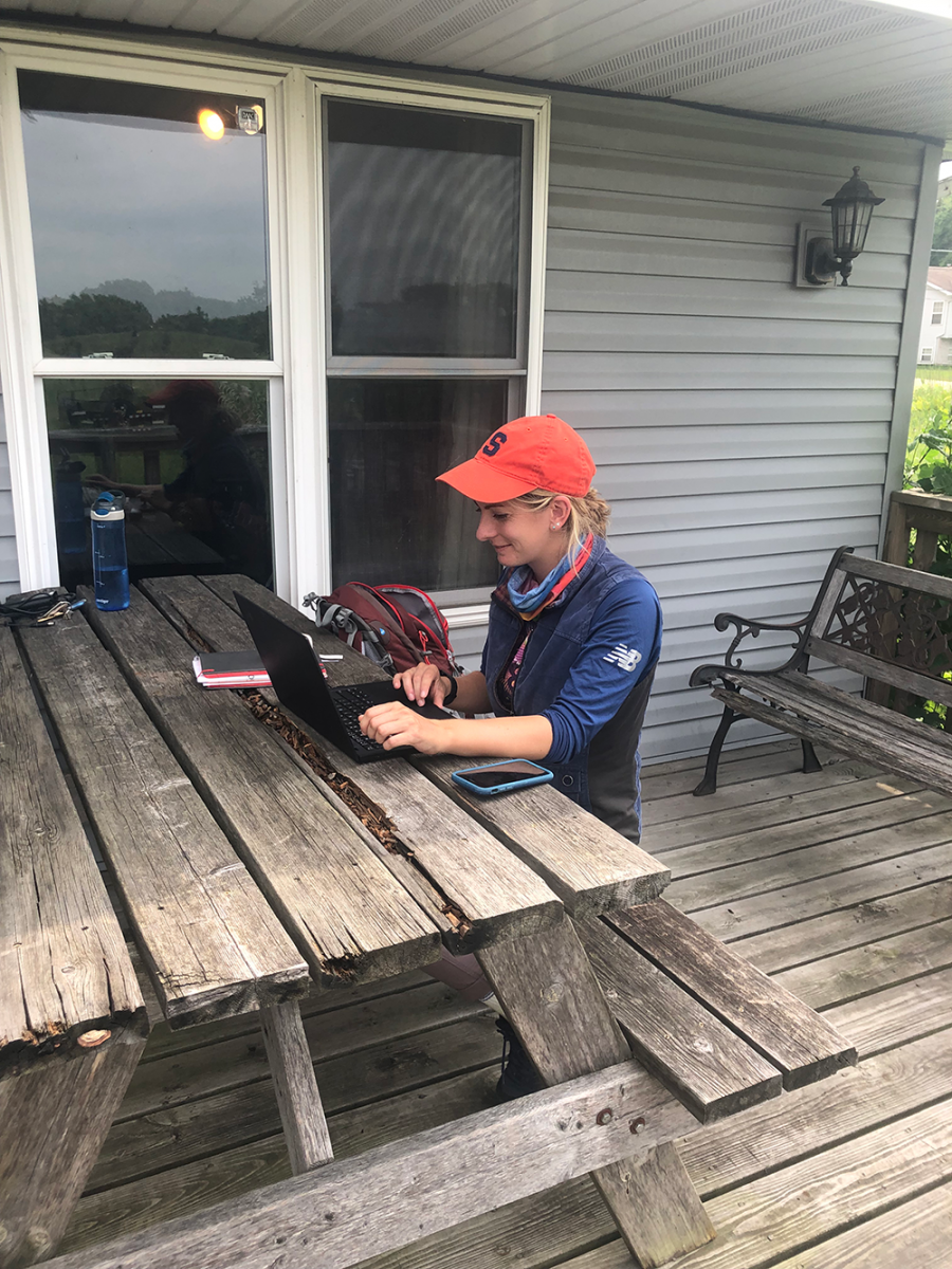 A woman in an orange cap sits at a picnic table on a porch while using a laptop computer.
