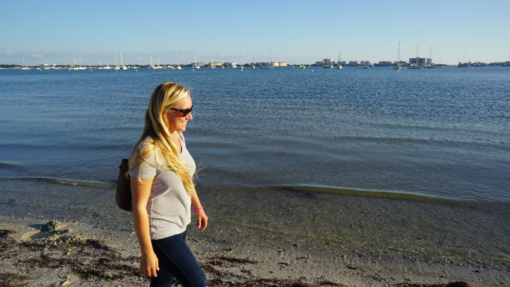 Sheila Scolaro walks on a beach along Tampa Bay. Across the water, moored sail boats and building line the shore.