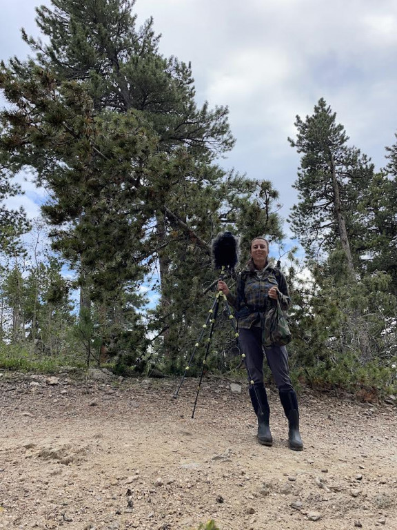 Acoustic ecologist, Laura Giannone stands in front of some trees holding a microphone attached to a long tripod. 