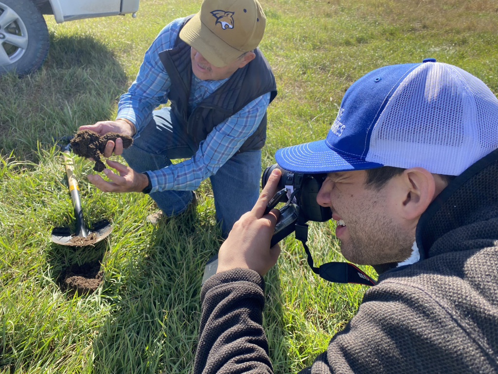 Mark Kossler kneels above a dug up hole in the grass, holding a clump of soil in his hands.