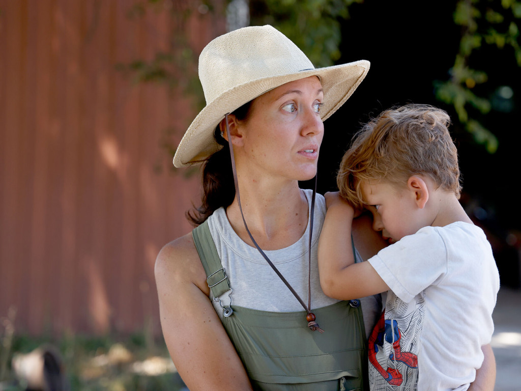 A woman in a large brimmed hat and overalls carries a young child who looks sleepy.
