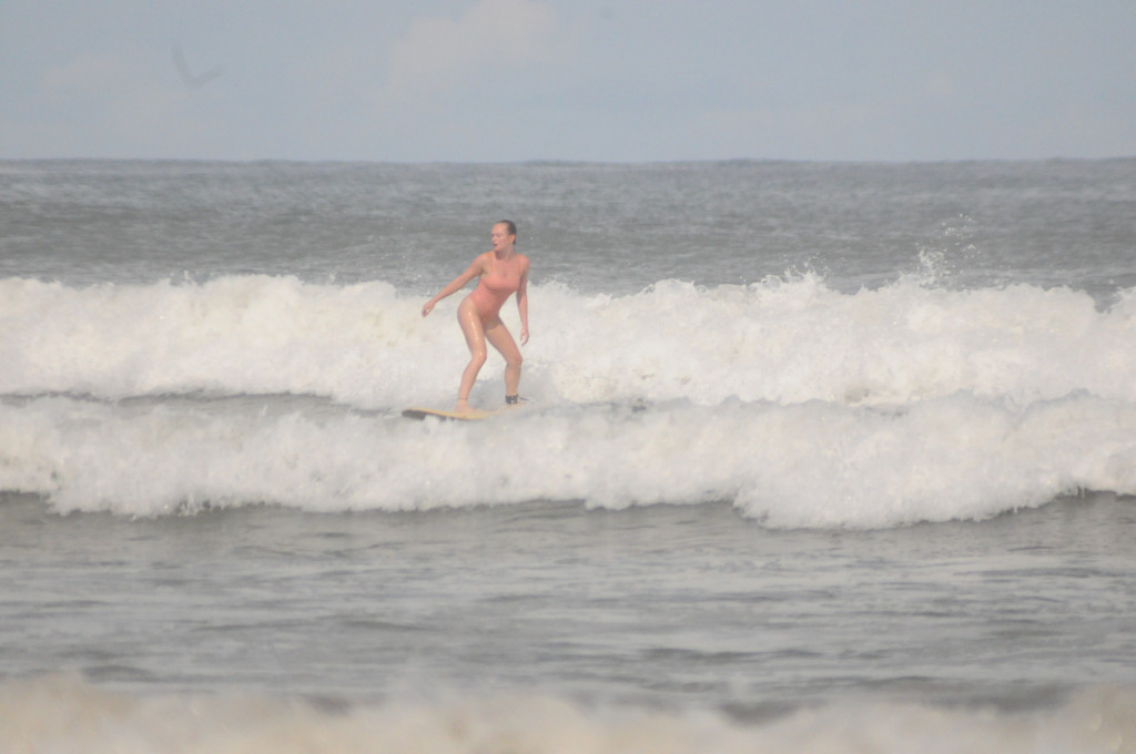 The author stands on a surfboard while riding a wave. A wall of ocean foam pushes her forward.
