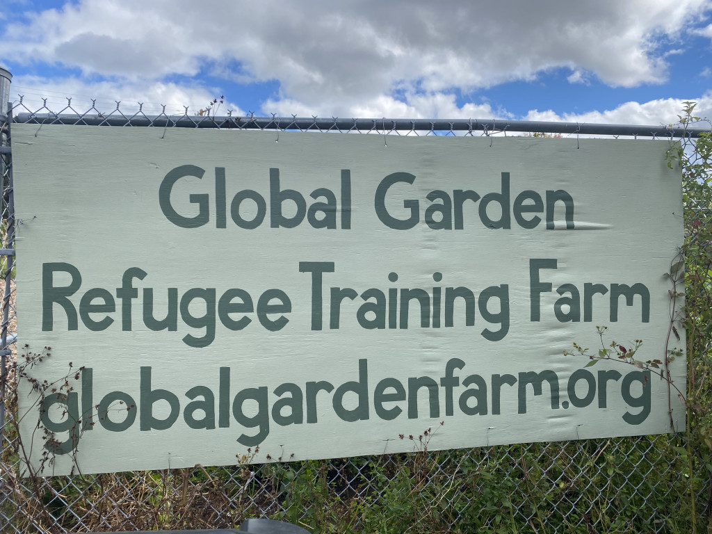 A sign hangs on a chain-link fence, reading "Global Garden Refugee Training Farm."