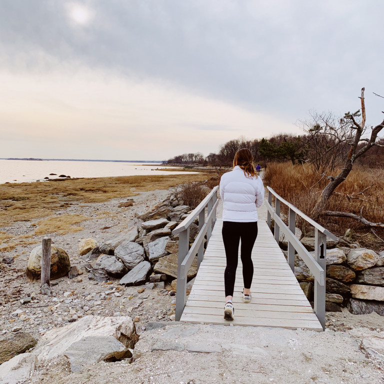 A woman in a white jacket walks away from the camera on a small wooden bridge surrounded by wintery brush and the Long Island Sound in the distance.