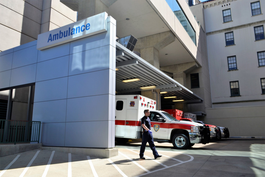 The exterior of an emergency room. An ambulance is parked outside and an man in an EMT uniform walks passed it.