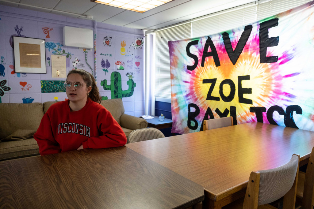 A person in a red shirt sits at a table. A colorful banner in the window reads "Save Zoe Bayliss."