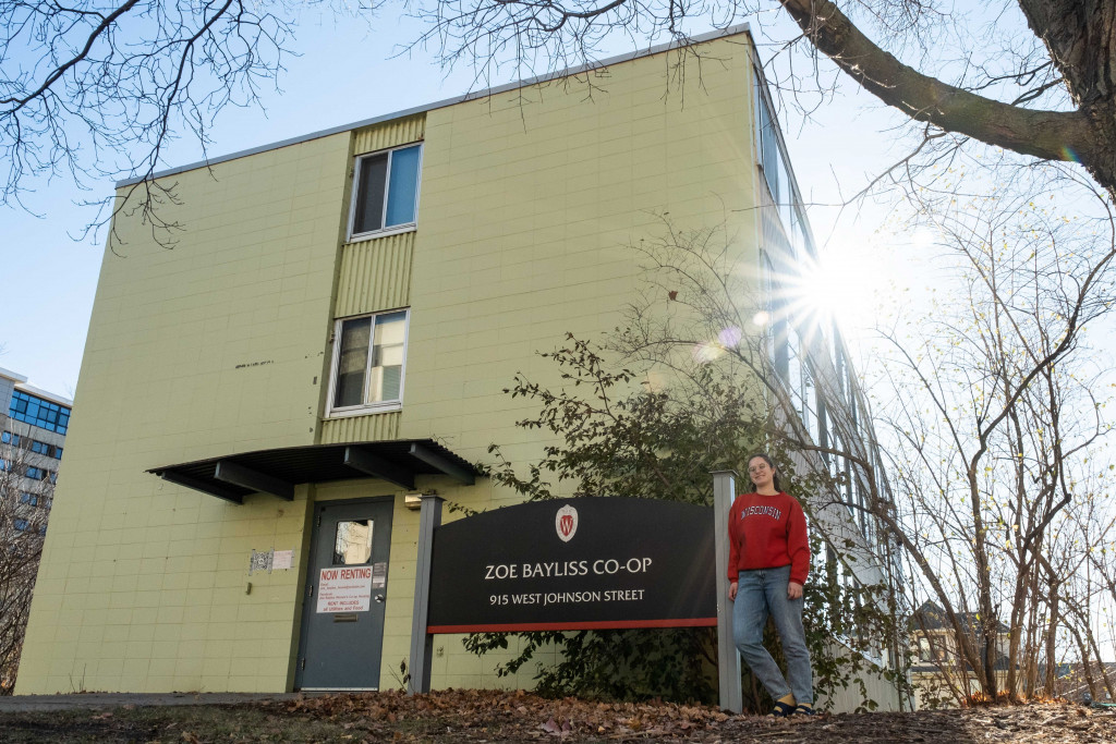 A person in a red shirt stands in front of a light green building with a sign reading "Zoe Bayliss Co-op."