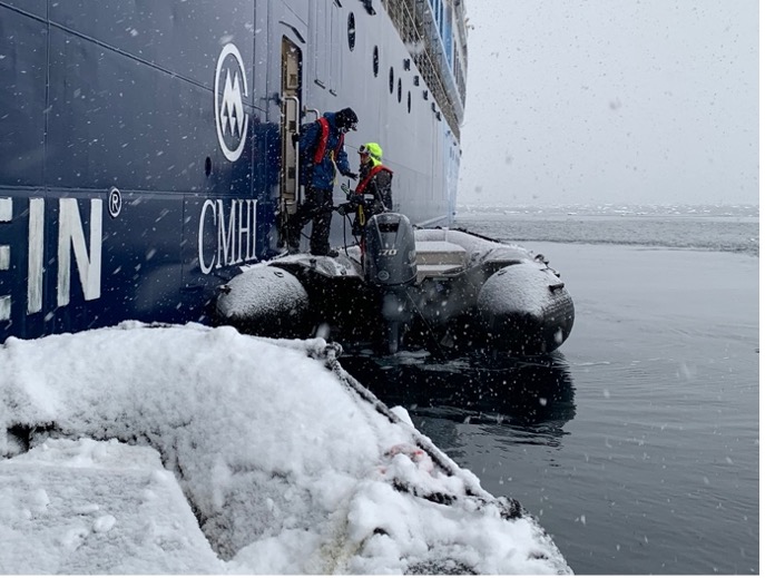 Two people step from a large ship onto a small boat in the icy waters of Antarctica